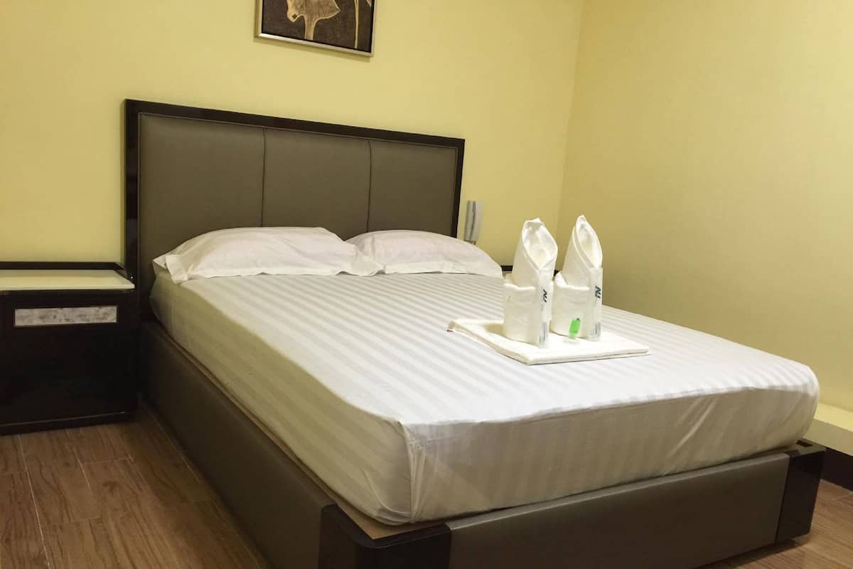 A neatly made bed with white linens, a dark headboard, and a tray with towels and a green bottle on the foot of the bed.