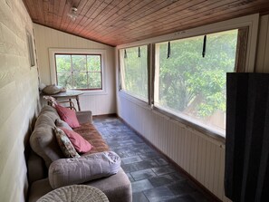 A cozy room with a wooden ceiling, a cushioned bench with pillows, a small table, and a window with a view of greenery.