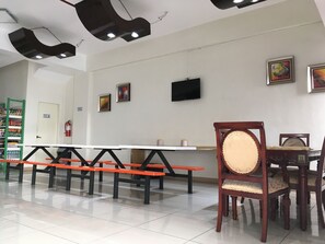 A dining area with a long white table and orange benches, wooden chairs with cushioned seats, and a television mounted on the wall. - Meaco Hotel - Valenzuela (Valenzuela)