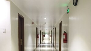 Un long couloir bien éclairé, avec des portes de chaque côté, un extincteur sur le côté droit et une horloge au mur.