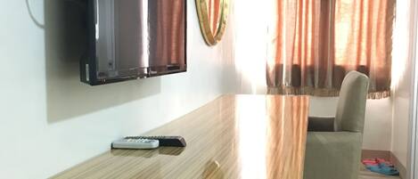 A modern living room with a flat-screen TV mounted on the wall, a wooden coffee table, a remote control, and a window with curtains.