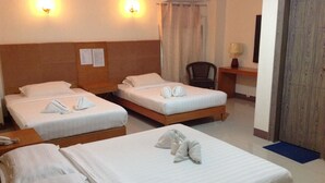 Une chambre d’hôtel avec trois lits simples, chacun agrémenté de serviettes soigneusement pliées. On y trouve deux lampes fixées au mur, une chaise, un bureau avec une lampe et une porte.