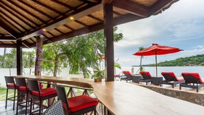 A covered outdoor seating area with a wooden table and chairs, red cushions, and a view of the sea. - The Bridge Huts (Ceningan Island)