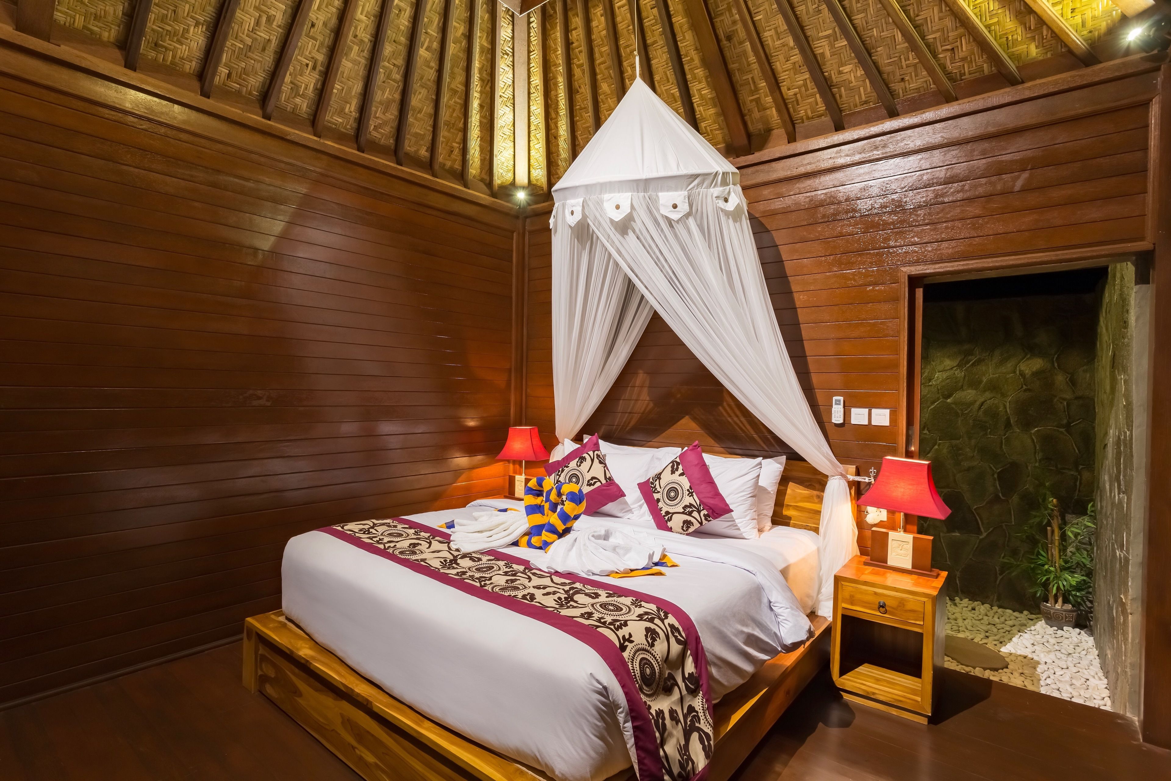 A wooden room with a bed, two nightstands, a red lamp, and a stone wall.