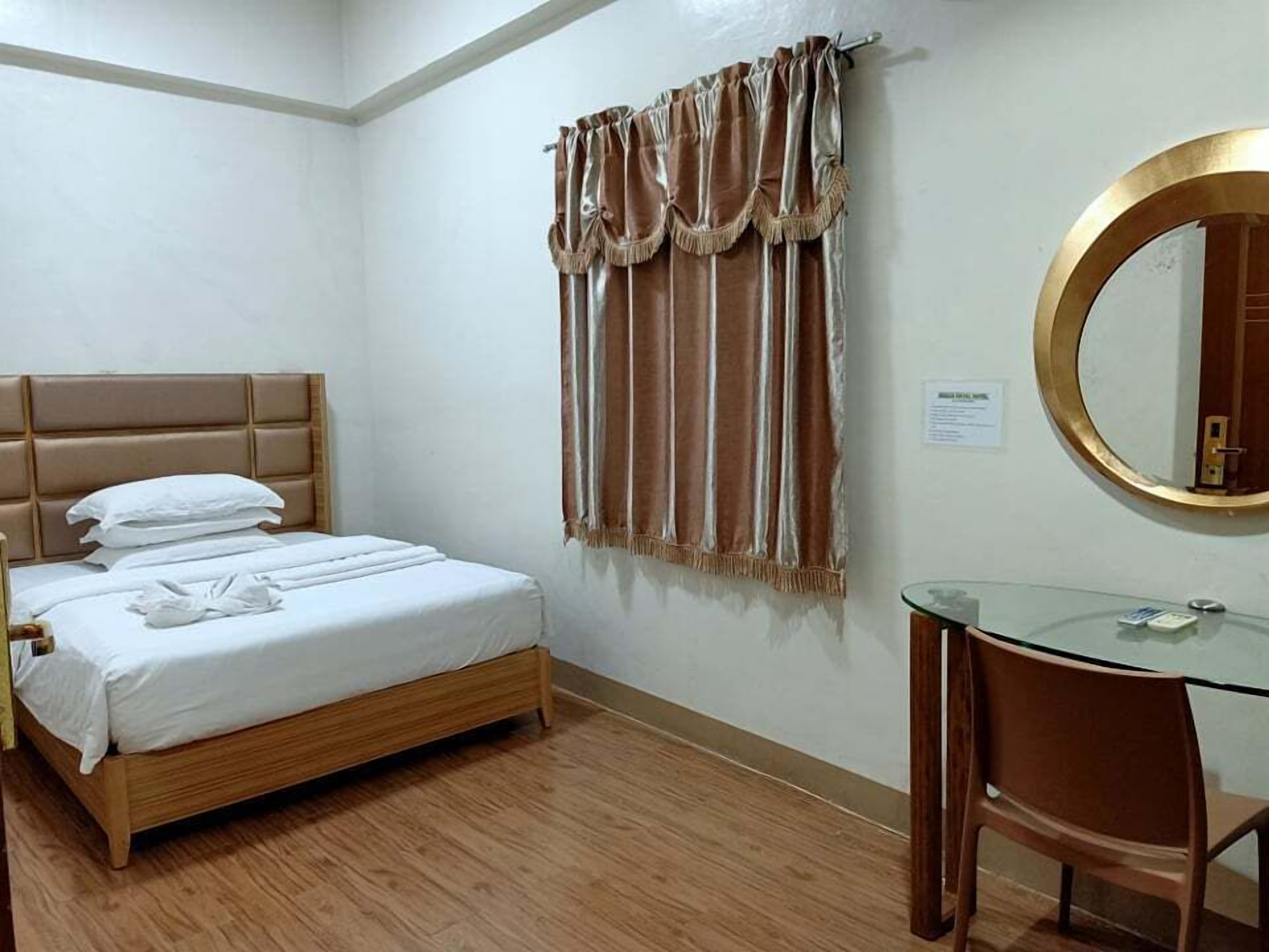 Habitación de hotel con una cama de madera, una mesa de vidrio, un espejo redondo y una ventana con cortinas.