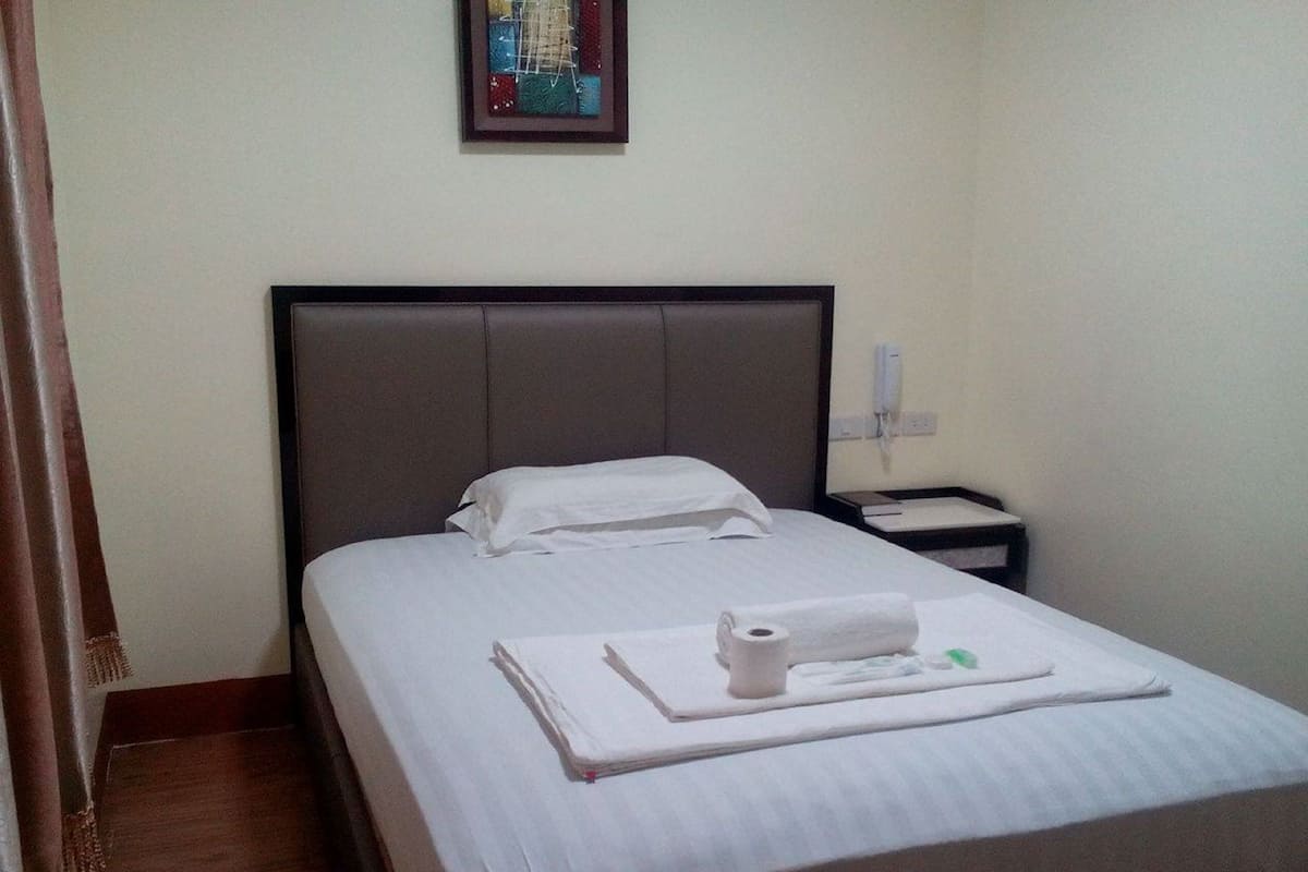 Een keurig opgemaakt bed met witte lakens, een kussen, een opgerolde handdoek en een klein dienblad met een flesje en zakdoekje.