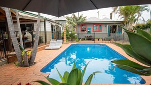 A rectangular swimming pool with a brick patio, surrounded by a fence, and a house with a red window frame in the background. - Backpackers Newcastle (Hamilton East)