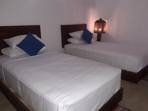 Two single beds with white linens and blue pillows, a nightstand with a lamp, and a plain white wall. - Ladun Sigiri Villa (Sigiriya)