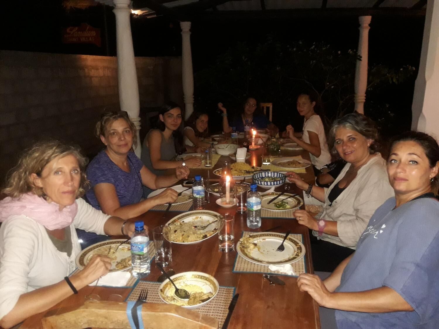 A group of people dining at an outdoor table with candles, plates of food, and water bottles.
