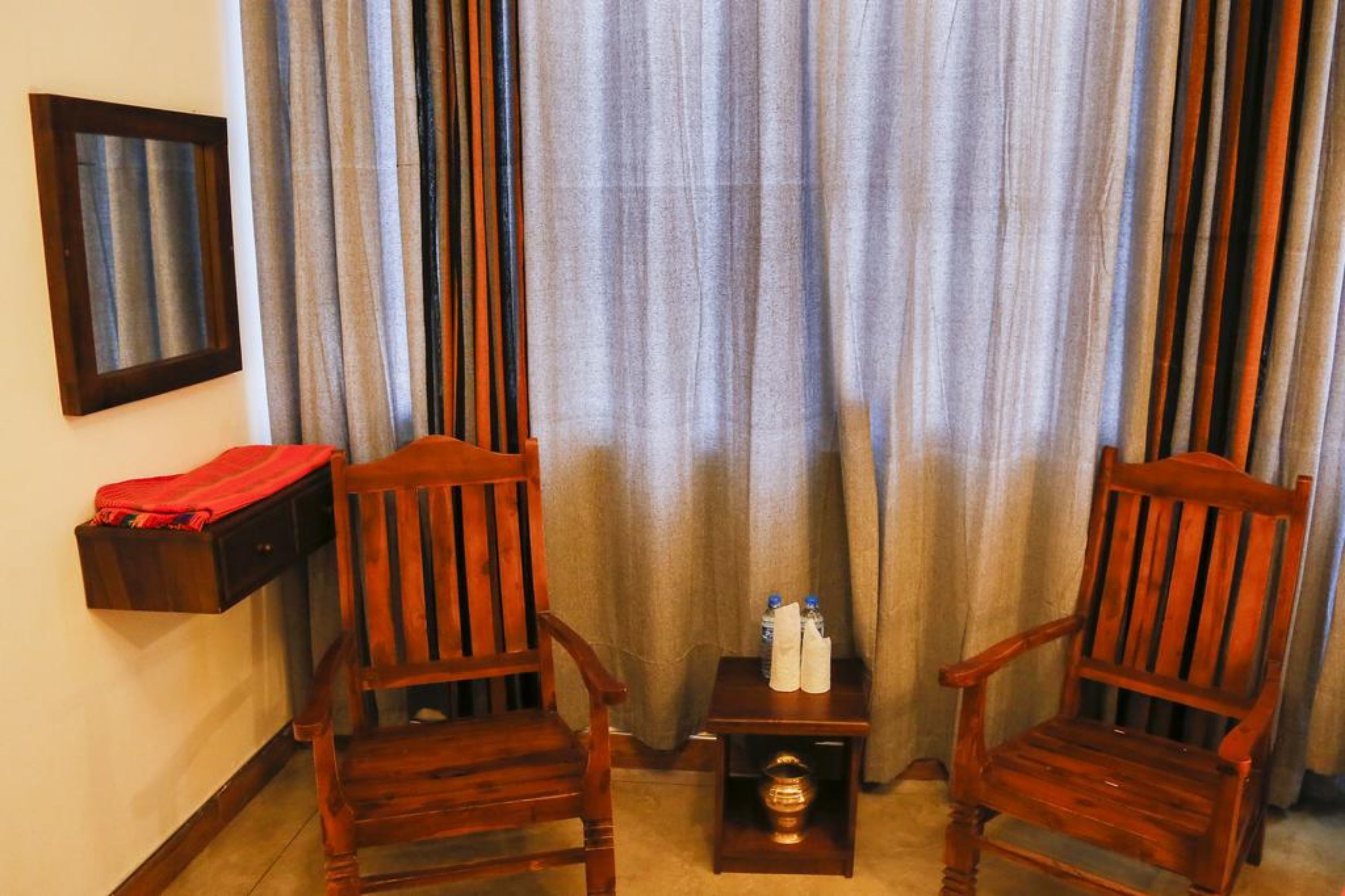 Una habitación con dos sillas de madera, una mesita con botellas de agua y un paño rojo sobre una estantería.