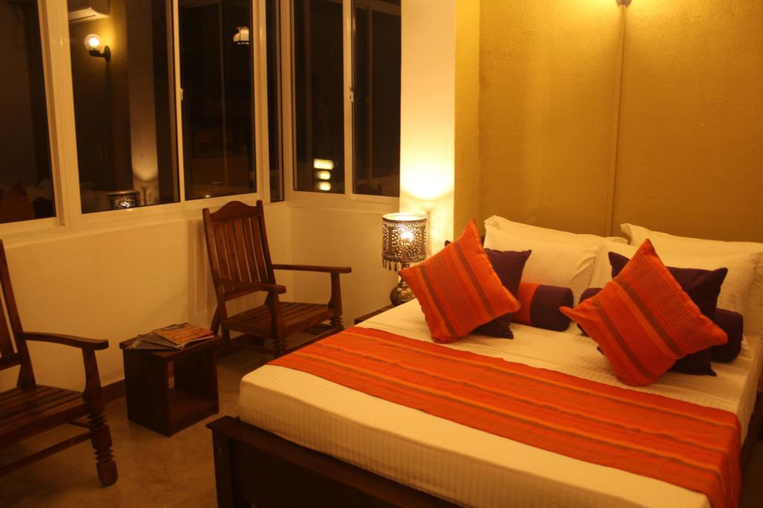 A neatly made bed with a striped blanket and multiple pillows, a wooden chair, a small table with a lamp, and a window with a view of the outside.