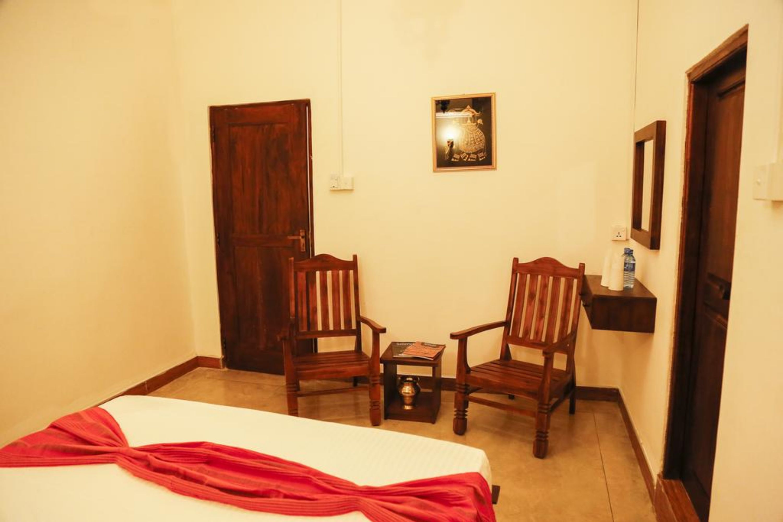 A room with a bed, two wooden chairs, a small table, a wall-mounted shelf with a bottle, and a door.