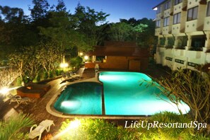 A swimming pool with lounge chairs and a small building, surrounded by trees and a multi-story building in the background. - Life Up Resort and Spa (San Sai)