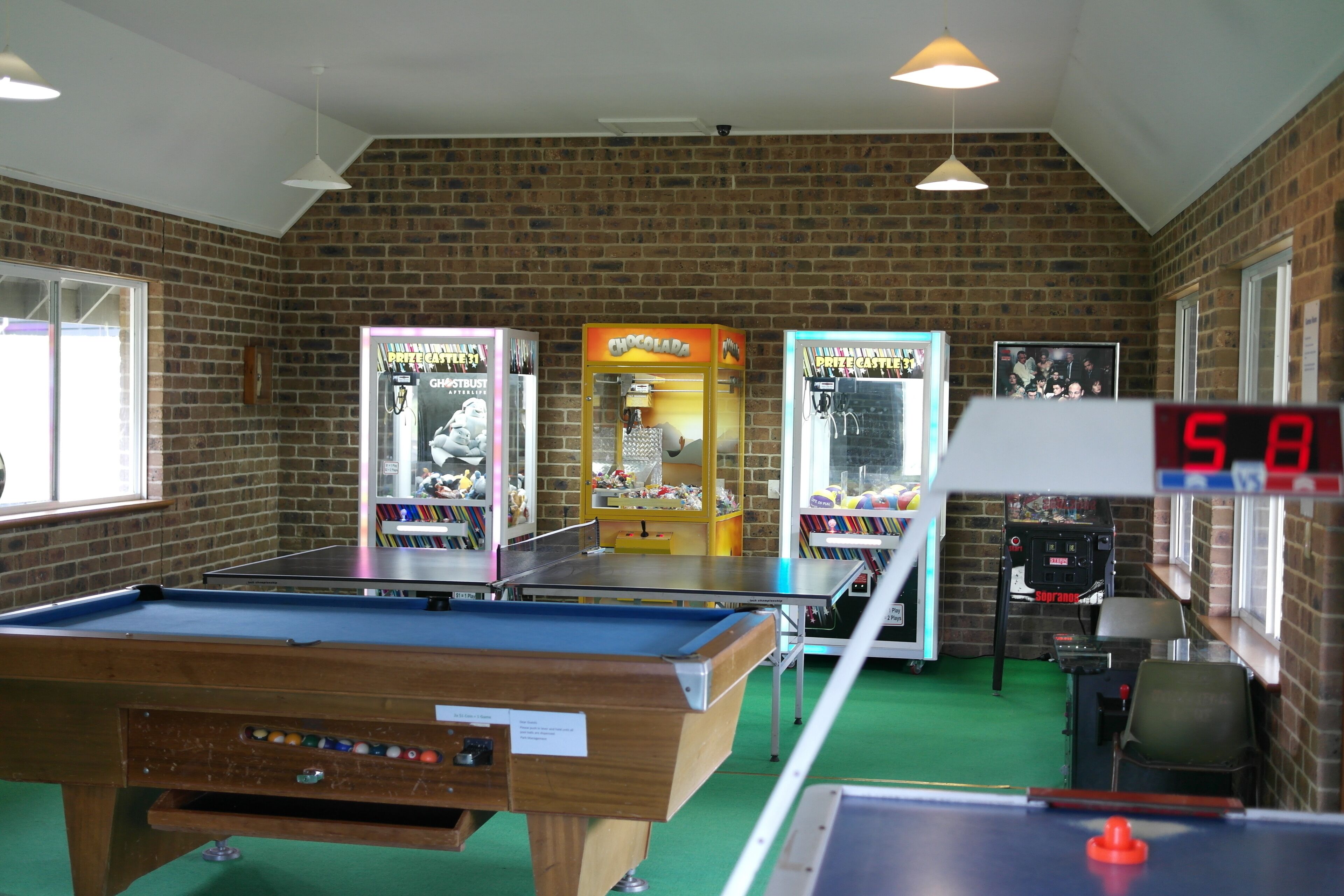 Une table de billard, des jeux d’arcade et une horloge numérique dans une pièce aux murs de briques et aux fenêtres.