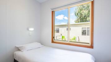A single-bed room with a large window, a bedside table, and a wall-mounted light.