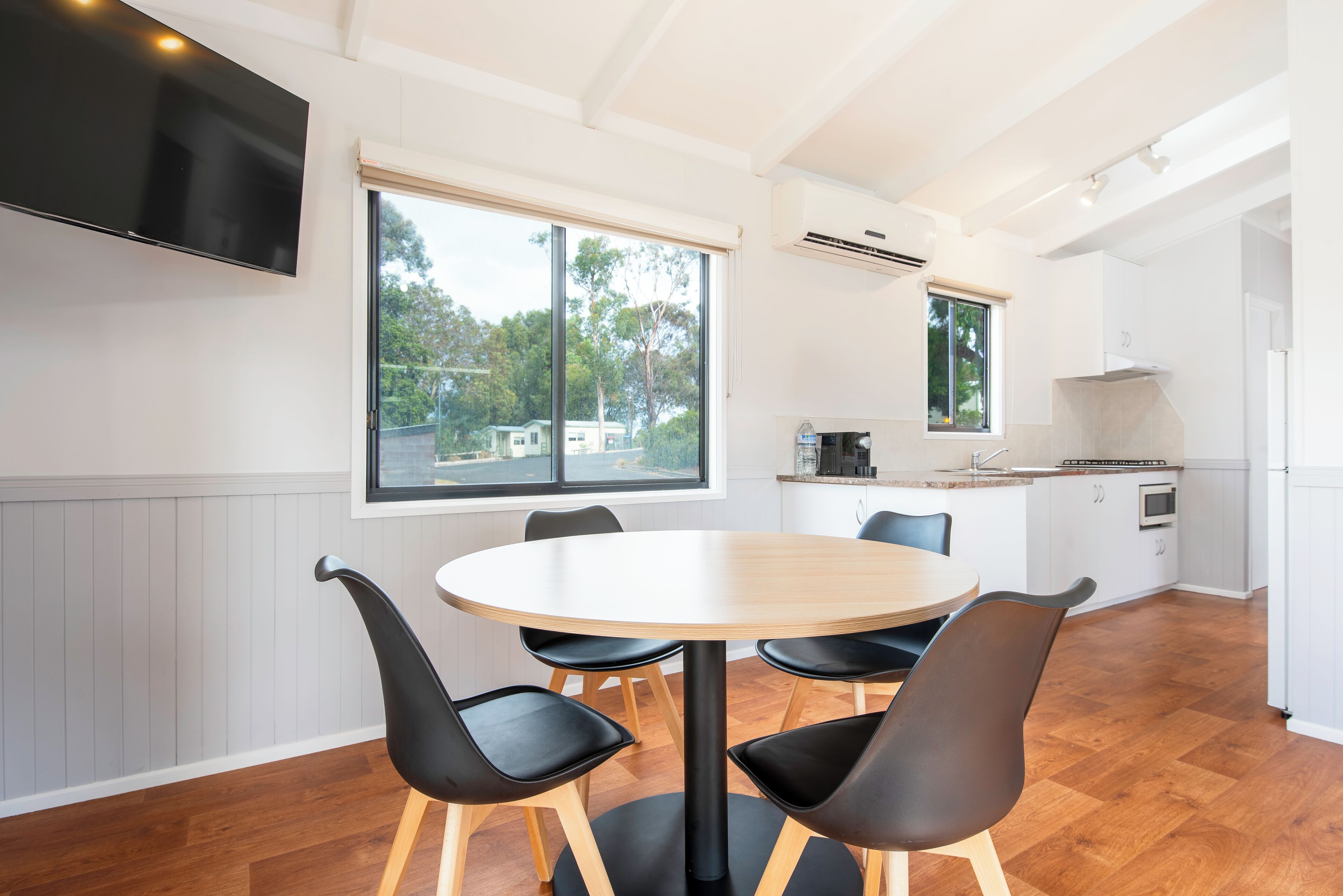Une cuisine moderne avec une table de salle à manger ronde et des chaises noires, de grandes fenêtres et un téléviseur à écran plat fixé au mur.