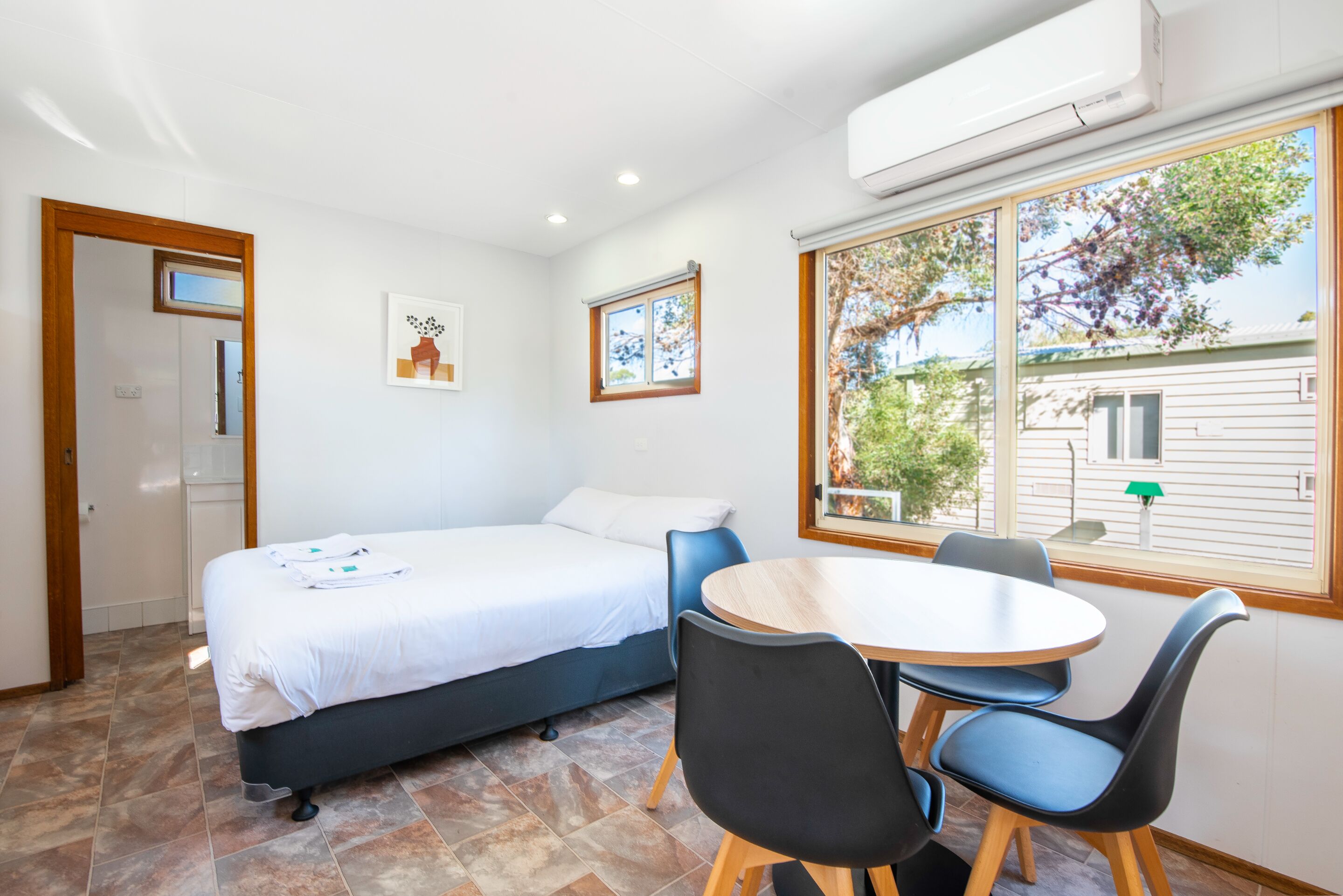 Ein Schlafzimmer mit Bett, einem Esstisch mit Stühlen, einem Fenster mit Blick auf Bäume und einer Tür.