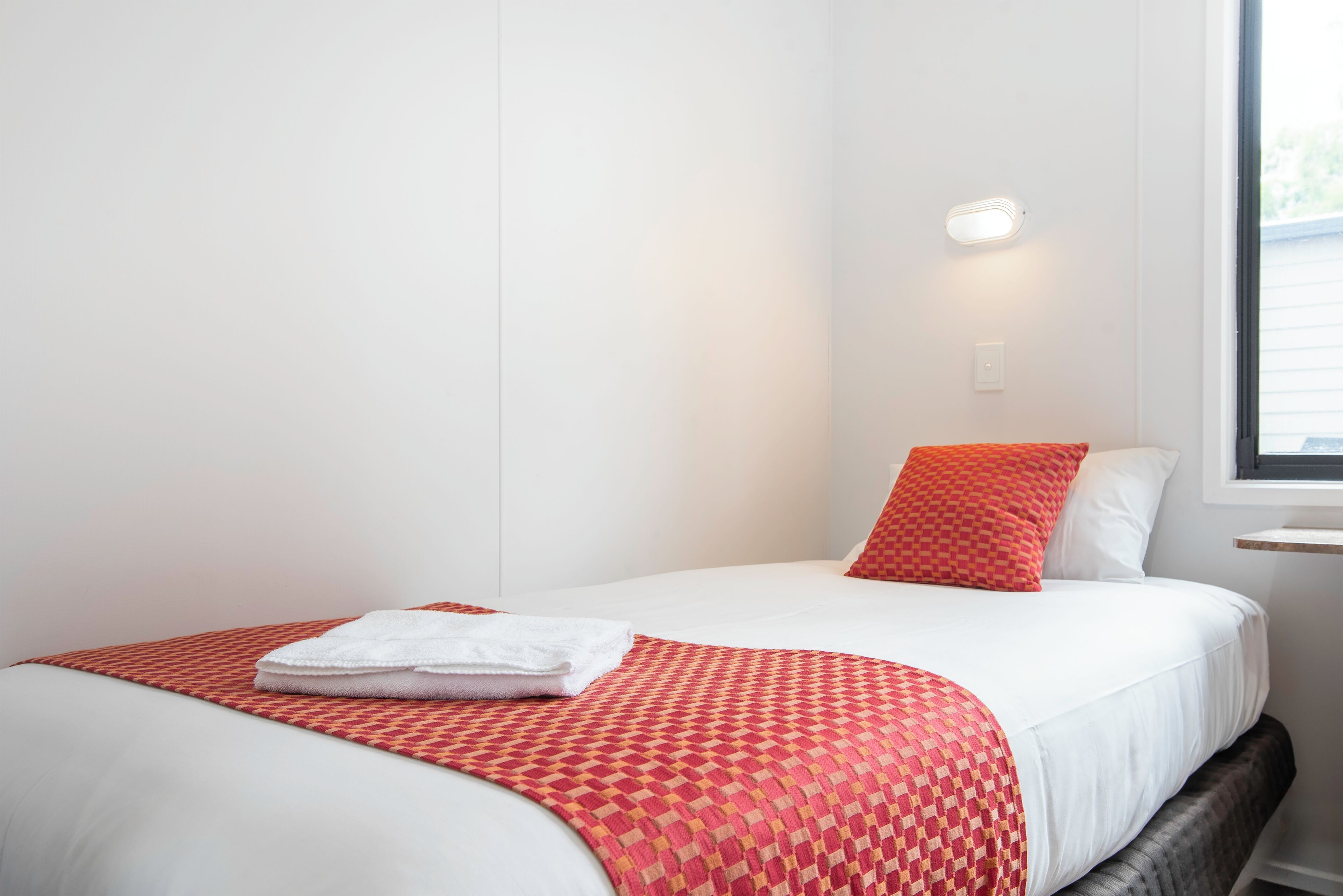 A neatly made bed with a patterned red blanket and a white pillow, a white wall, and a window with a view.