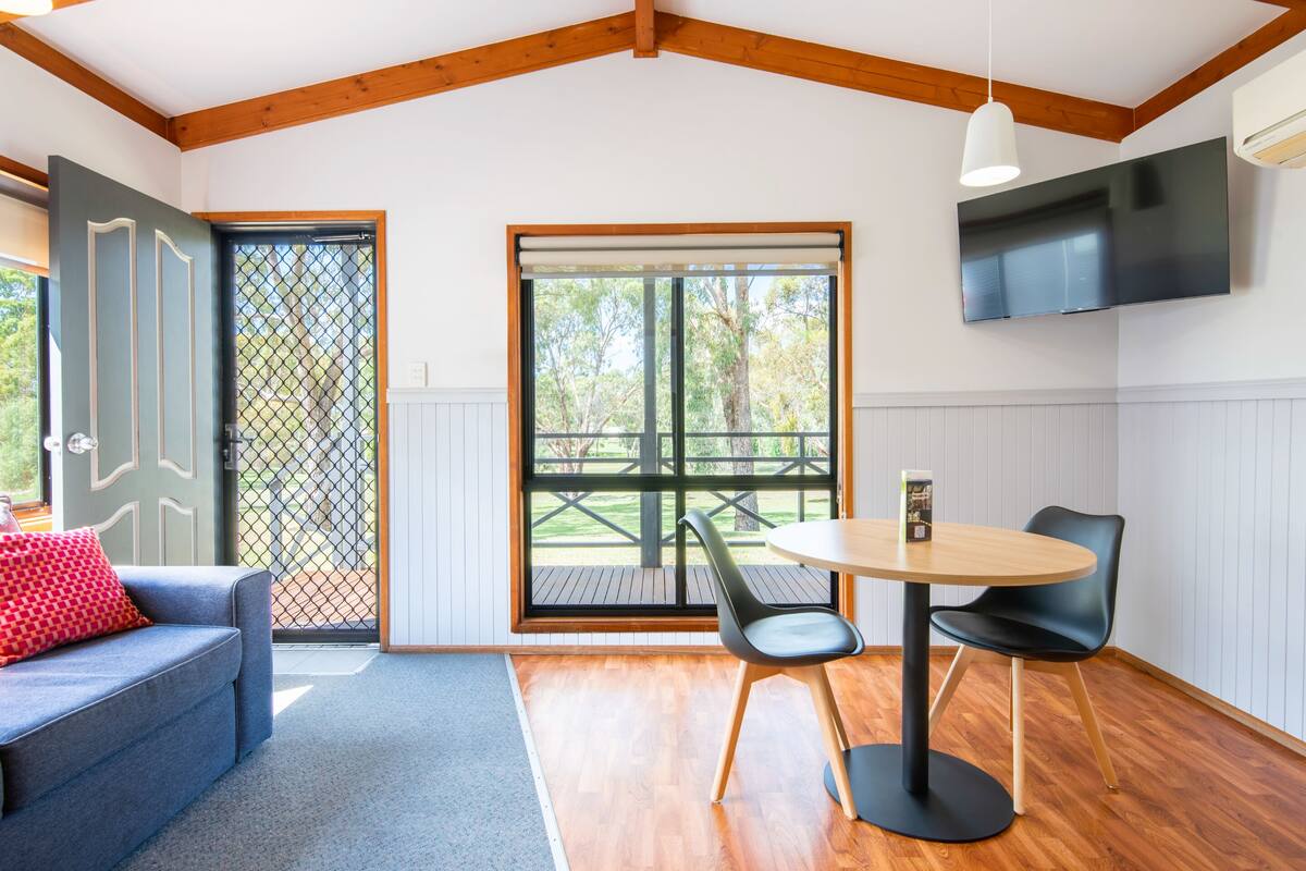 A modern interior with a wooden ceiling, a TV mounted on the wall, a round table with two chairs, a sofa with a red pillow, and a large window with a view of trees.