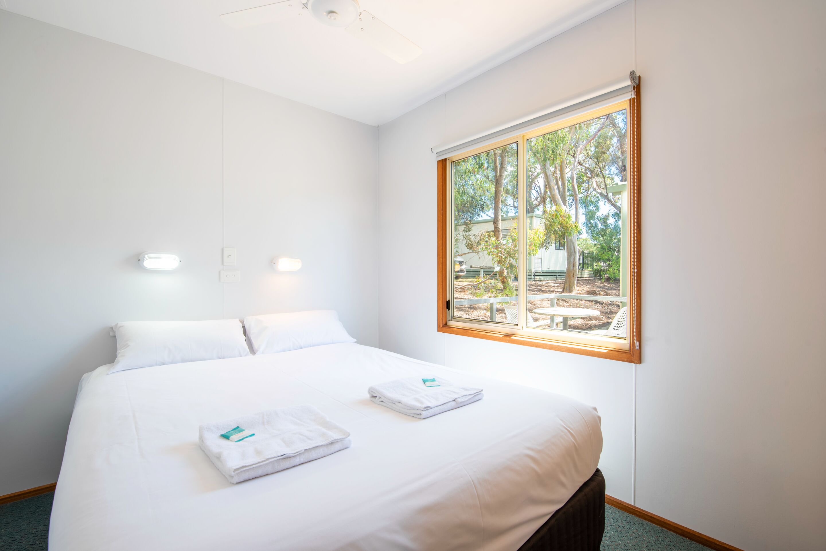 A hotel room with a large bed, two white pillows, a neatly made bed with towels, a window with a view of trees and a patio, and a ceiling fan.