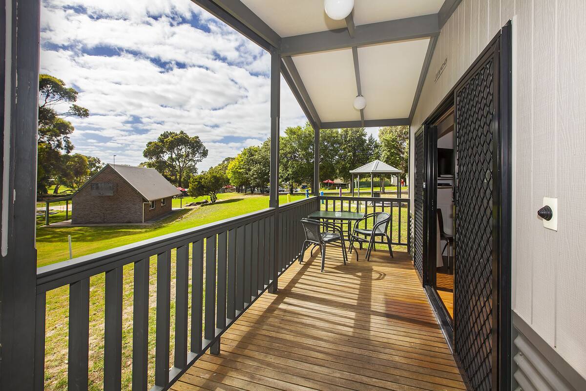 A covered outdoor area with a wooden deck, a table with chairs, and a view of a grassy field and a building in the distance.