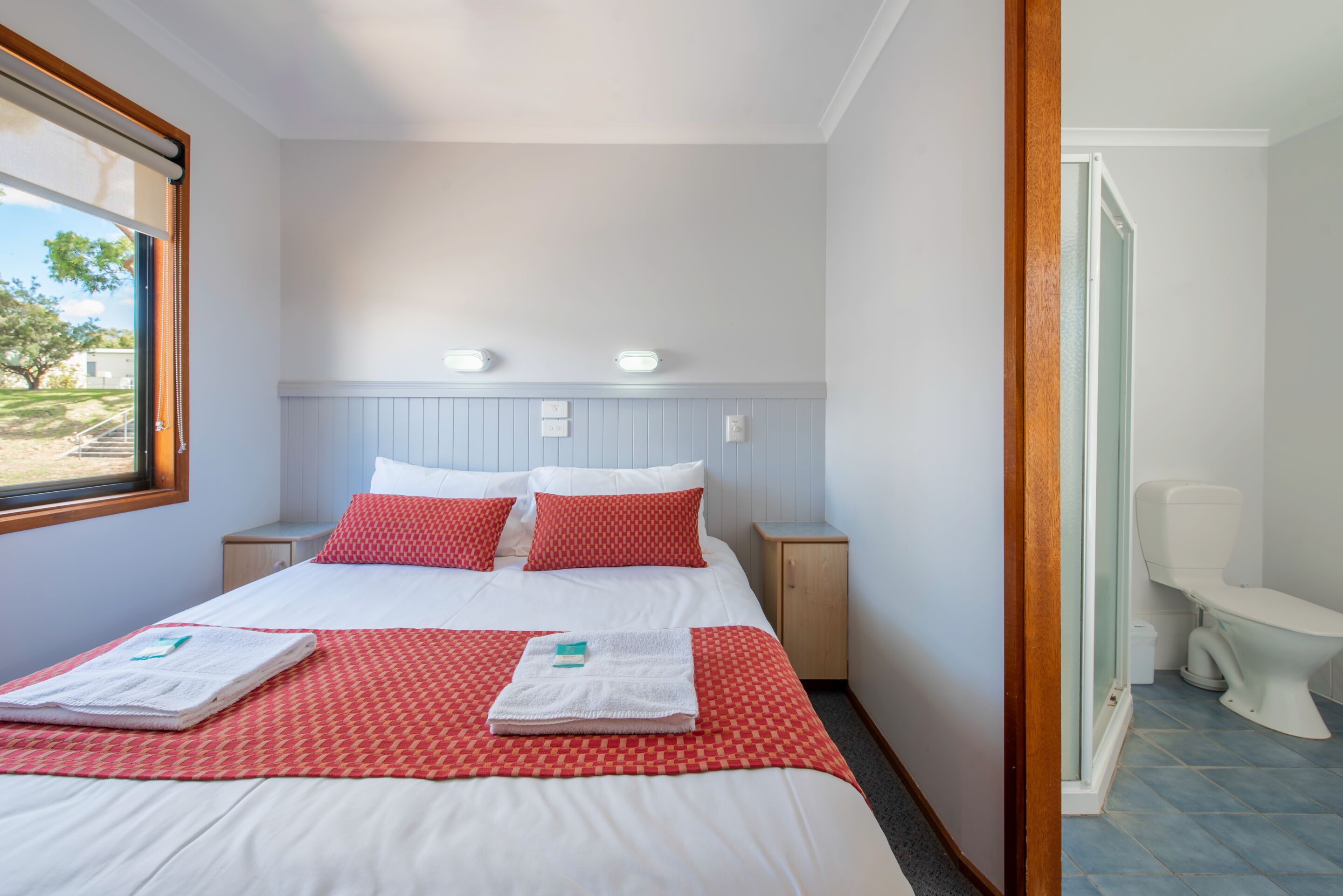 A neatly made bed with red and white bedding, a small bedside table, and a window with blinds.