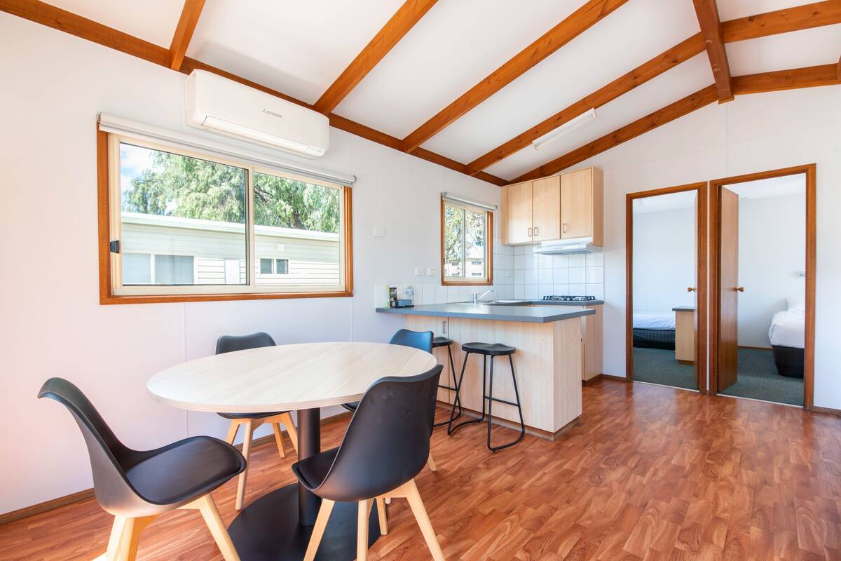 A modern kitchen with wooden beams, a dining area with a round table and chairs, and a view of a house through a window.