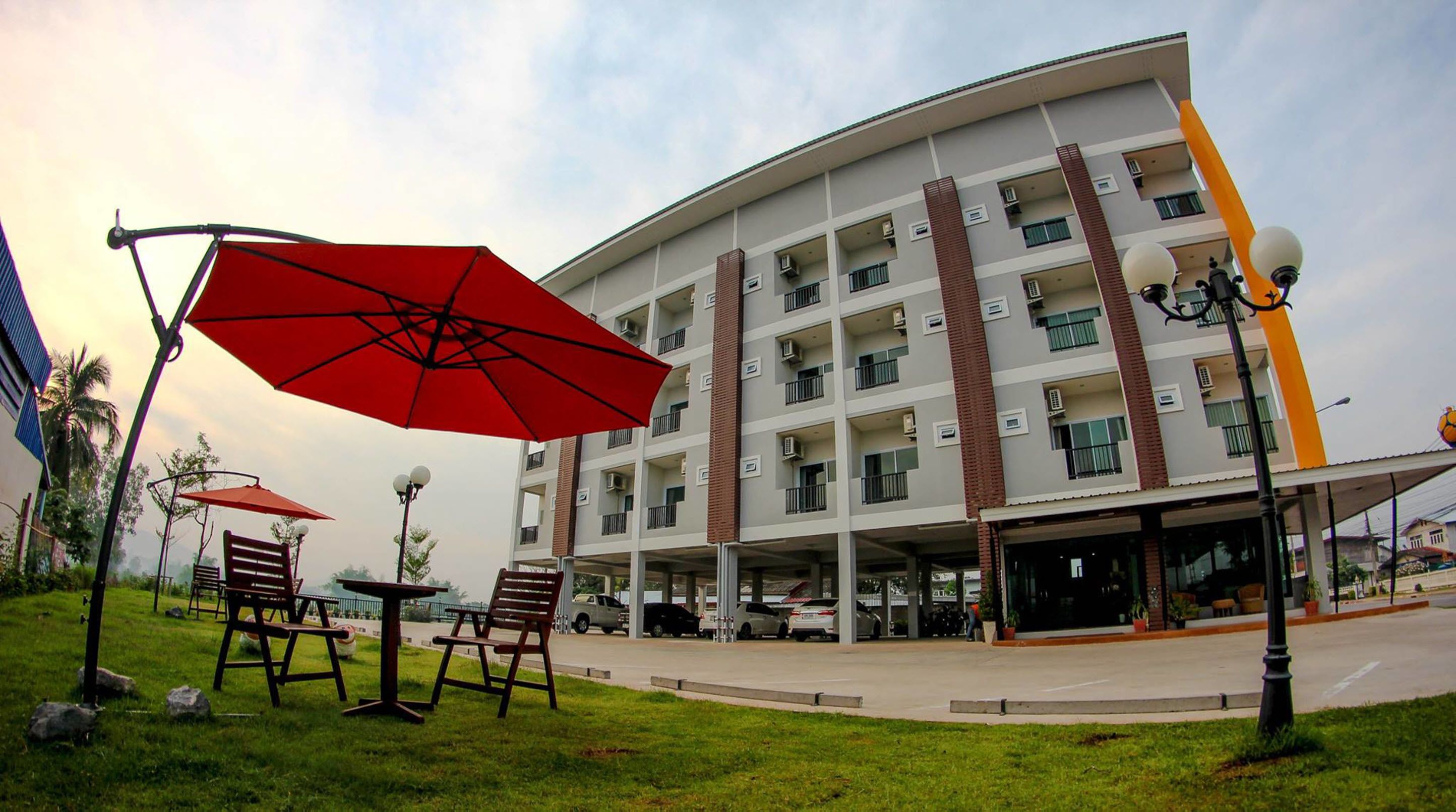 A multi-story hotel with a flat roof, balconies, and air conditioning units. There are outdoor seating areas with red umbrellas.