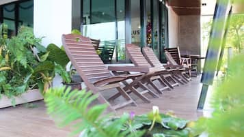 A row of wooden lounge chairs on a wooden deck with potted plants and a glass building in the background.
