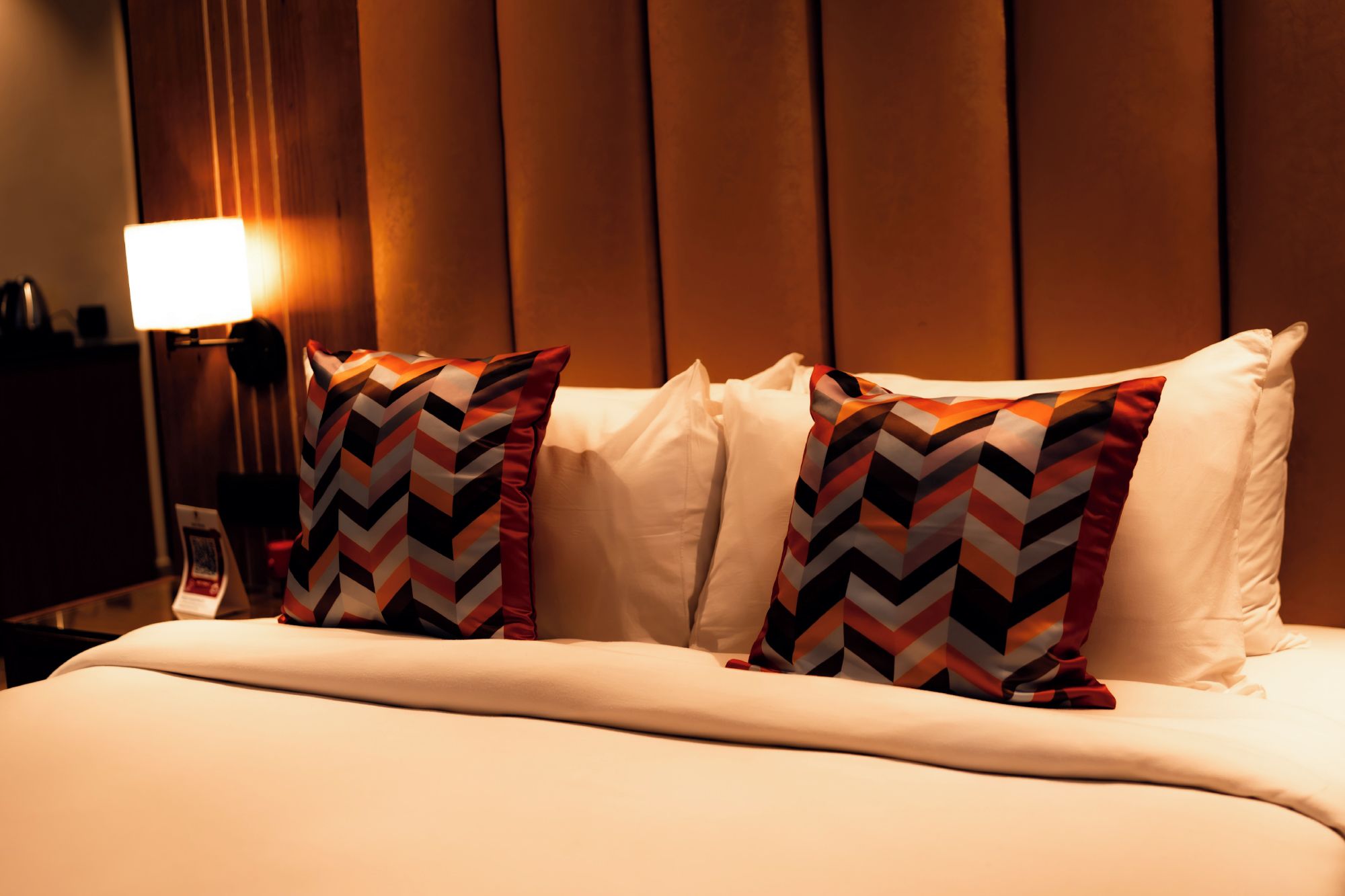 Habitación de hotel con cama, dos almohadas estampadas con diseño en zigzag, lámpara de noche y pared con paneles verticales.