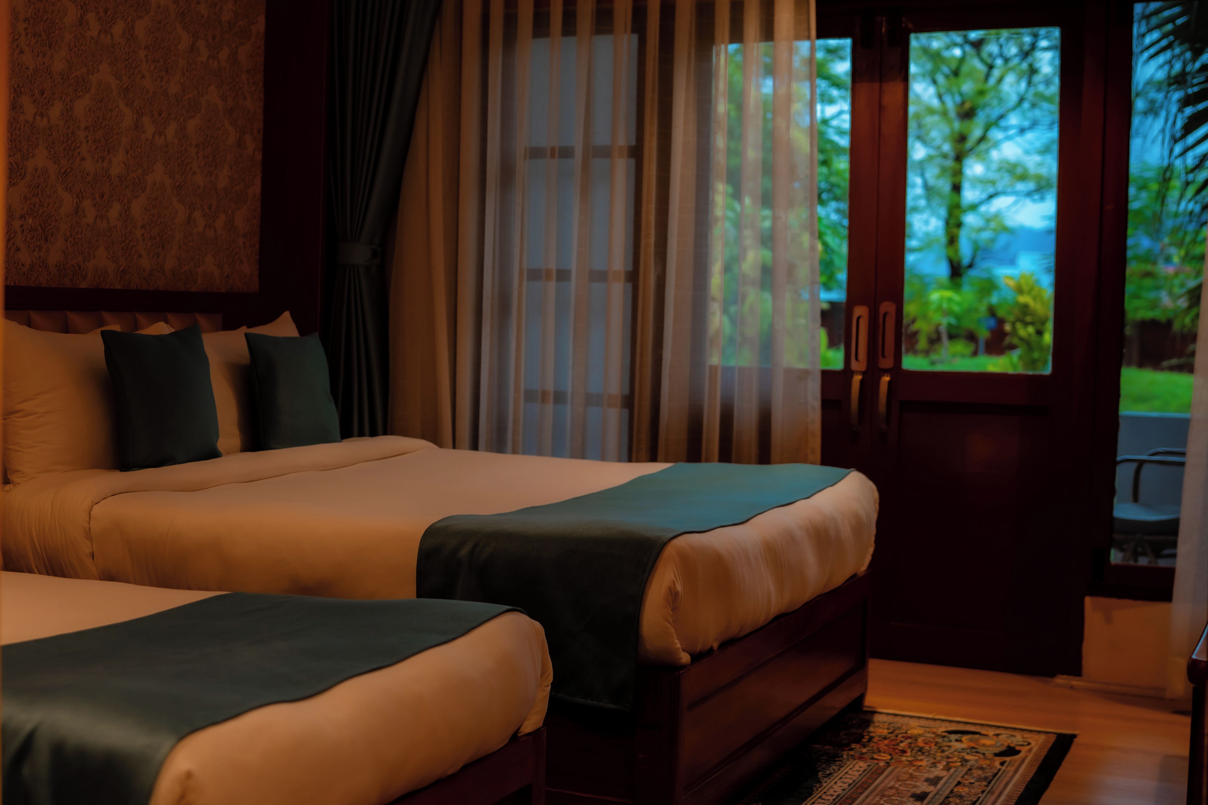 A hotel room with two beds, a view of greenery through the door, and a patterned rug.