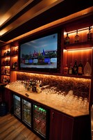Zona de bar con pantalla de televisión, estanterías con botellas de licor y un refrigerador con bebidas.