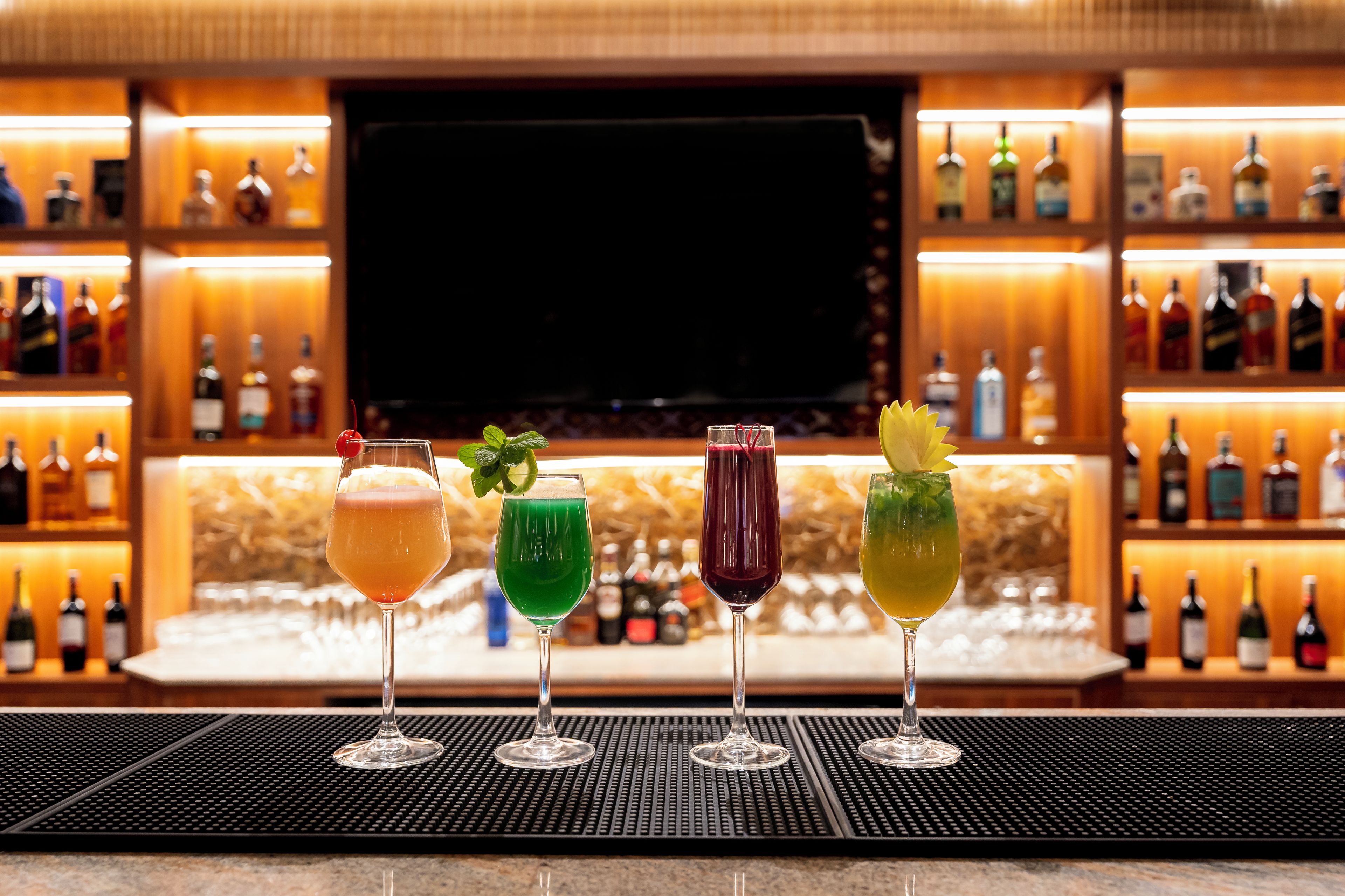 Zona de bar con tres vasos de bebidas de colores diferentes, una pantalla de televisión y estantes llenos de botellas al fondo.
