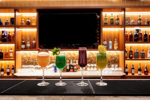 A bar area with three glasses of different colored drinks, a television screen, and shelves of bottles in the background. - Dreamland Gold Resort (Tilottama)