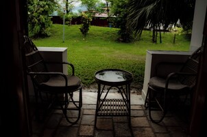 A patio with two wicker chairs and a small table, overlooking a grassy yard with trees and a building in the background.