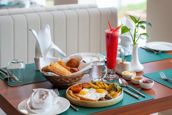 Desayuno con un plato de huevos, tostadas y una bebida roja, una cesta de bollería y un florero con plantas.