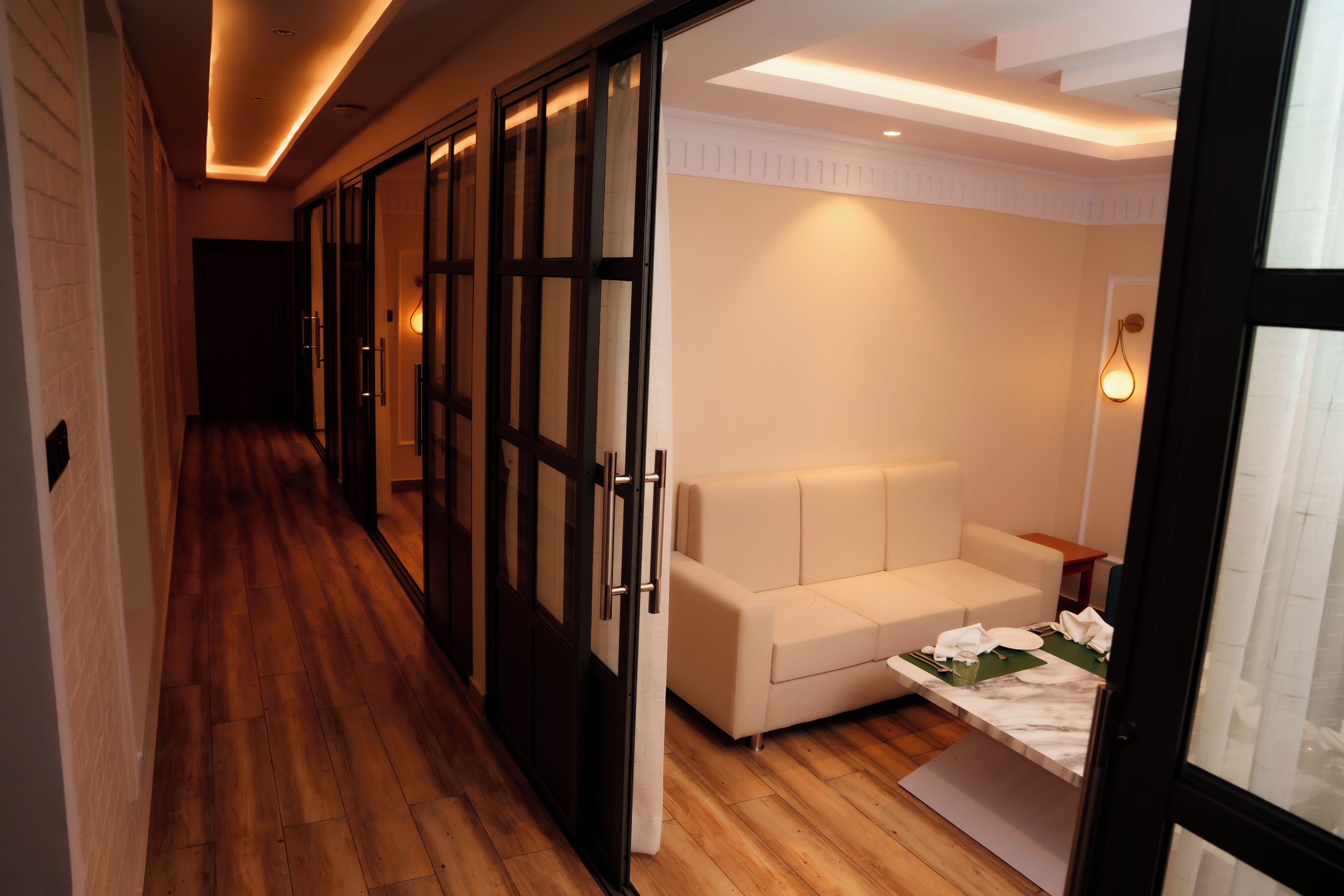 A hotel corridor with wooden flooring, a white sofa, and a dining area with a table set for two.