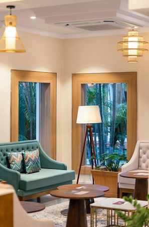 A modern hotel lobby with a sofa, armchair, floor lamp, and a view of tropical foliage through large windows. - Dreamland Gold Resort (Tilottama)