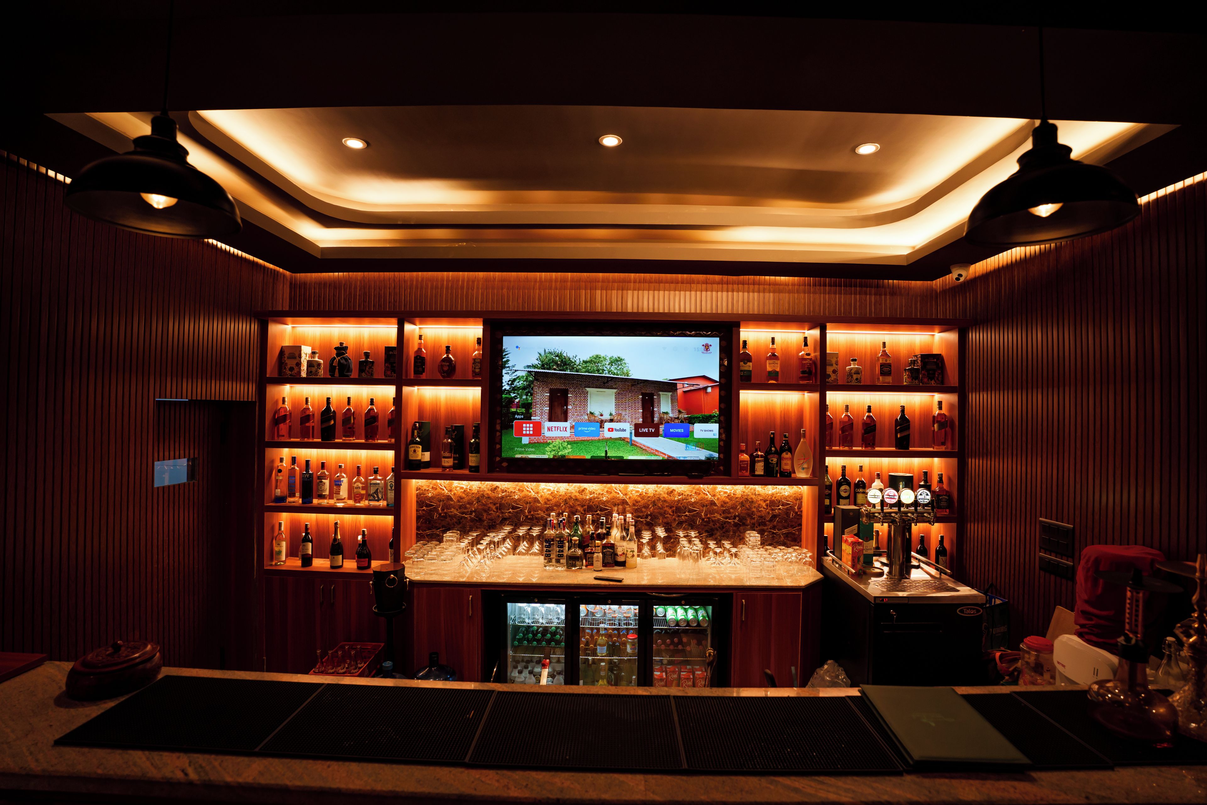 A bar area with shelves of liquor bottles, a television screen, and a bar counter with glasses and bottles.