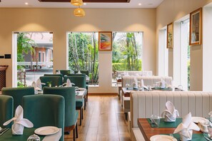 A restaurant with green upholstered chairs, white table settings, and large windows offering a view of the outdoors. - Dreamland Gold Resort (Tilottama)