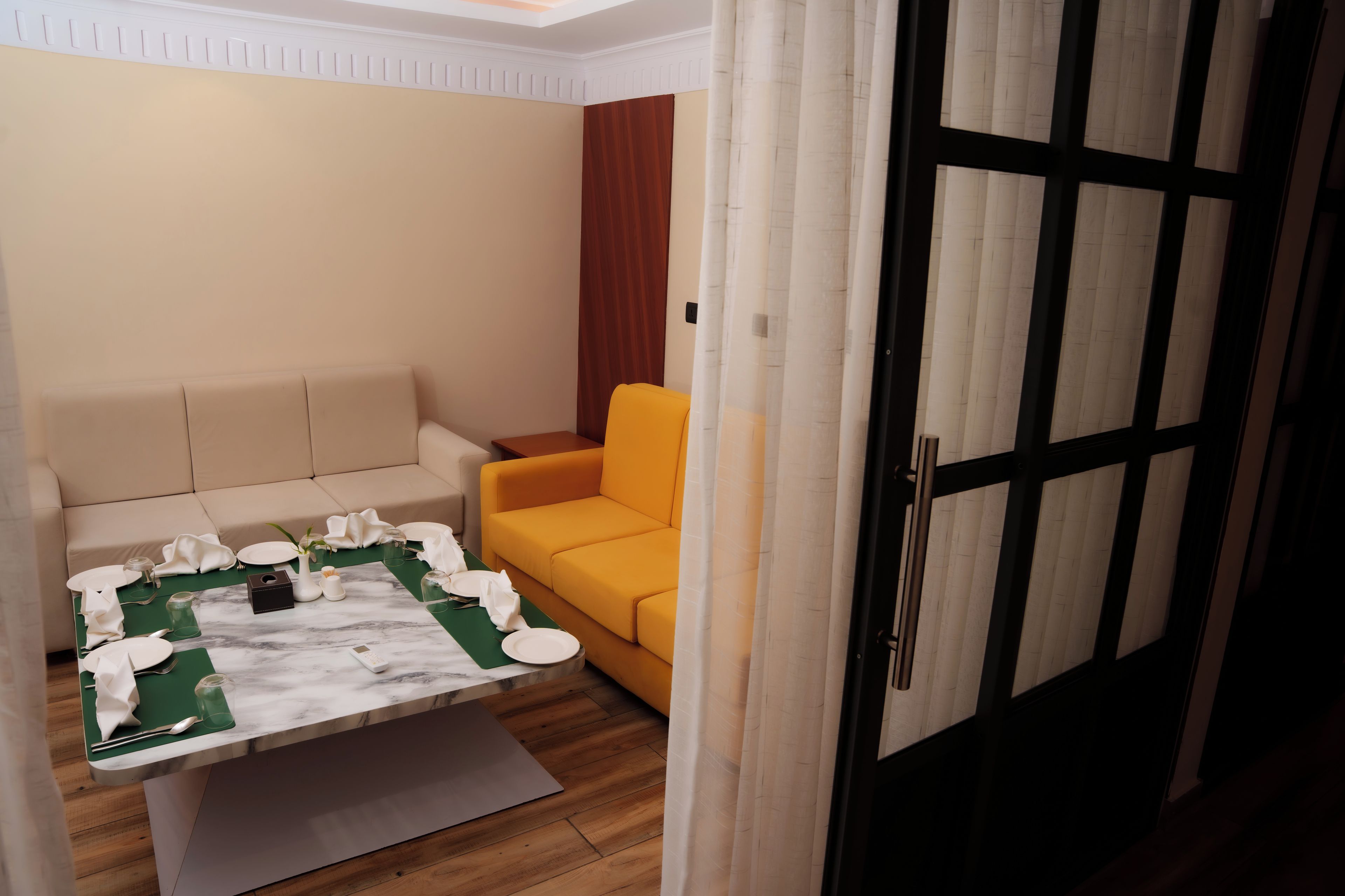 A room with a dining table set for four, a sofa, a yellow armchair, and a door with a black frame.