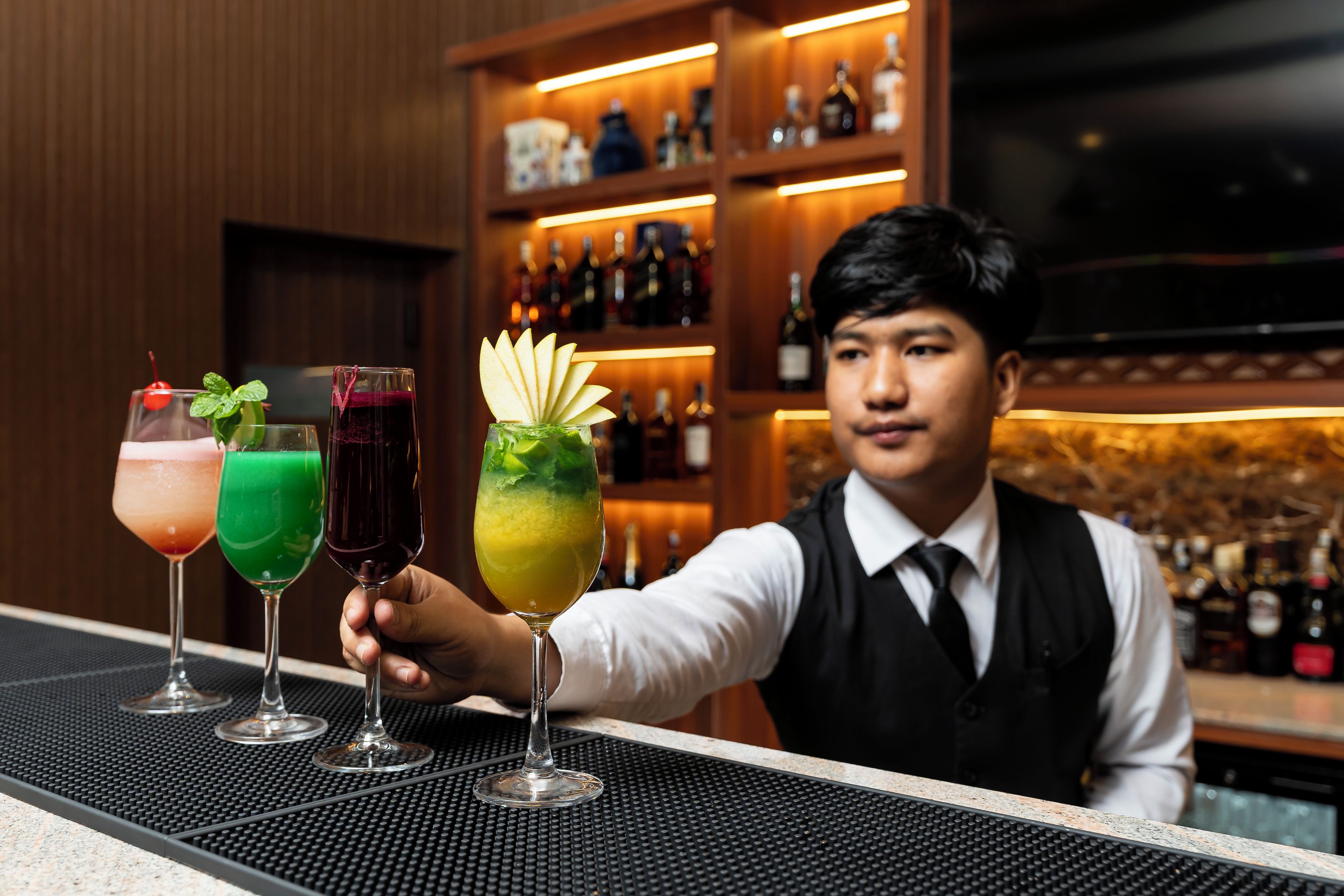 A bartender presenting three drinks at a bar with shelves of bottles in the background.