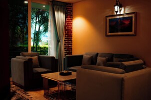 Un salón acogedor con sofá, sillones, una mesa de centro y vistas a la vegetación a través de una ventana con cortinas.