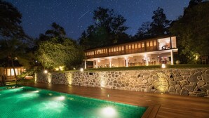 A swimming pool with a stone wall and wooden deck, surrounded by trees and a night sky with stars. - Kings Pavilion (Kandy)