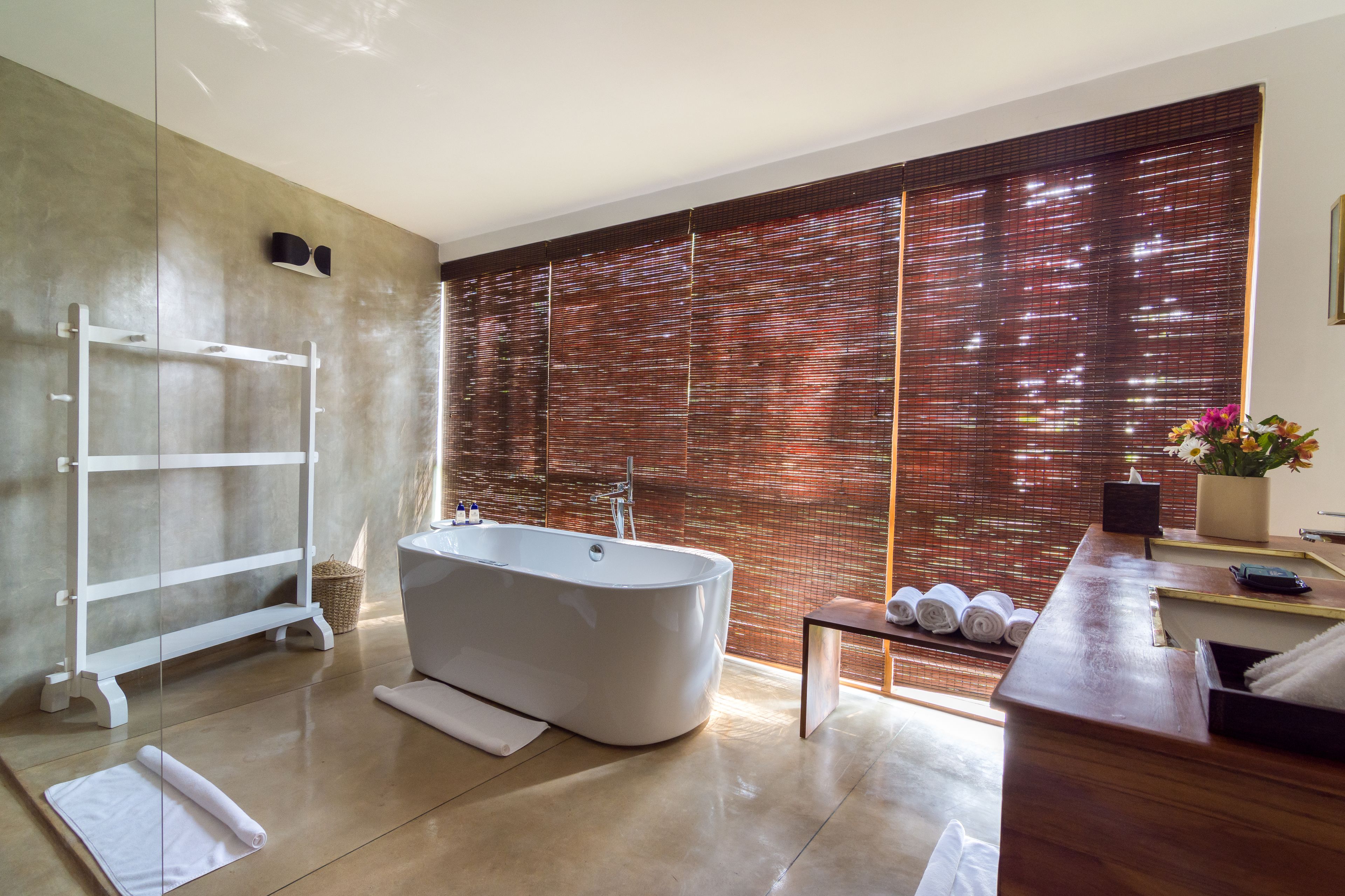 A modern bathroom with a freestanding bathtub, a wooden vanity, and a large window with a bamboo blind.