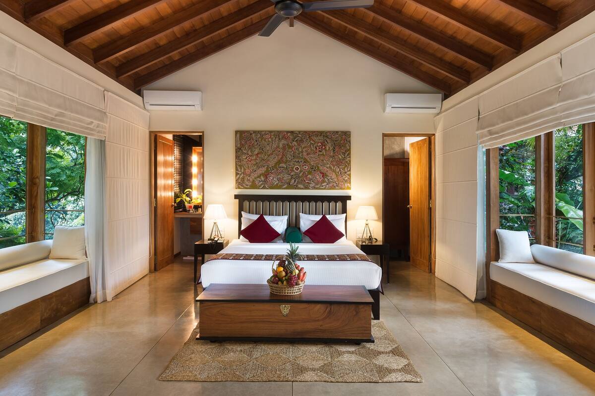 A spacious bedroom with a large bed, a wooden ceiling, and a view of greenery through the windows.