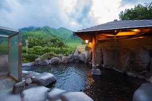 Un onsen japonais traditionnel, avec une structure en bois, un bassin bordé de pierres et une vue sur des collines verdoyantes en arrière-plan.