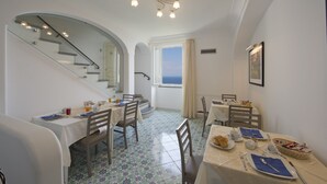 Uma área de refeições com uma escada que leva a uma varanda, paredes brancas e chão revestido de azulejos.