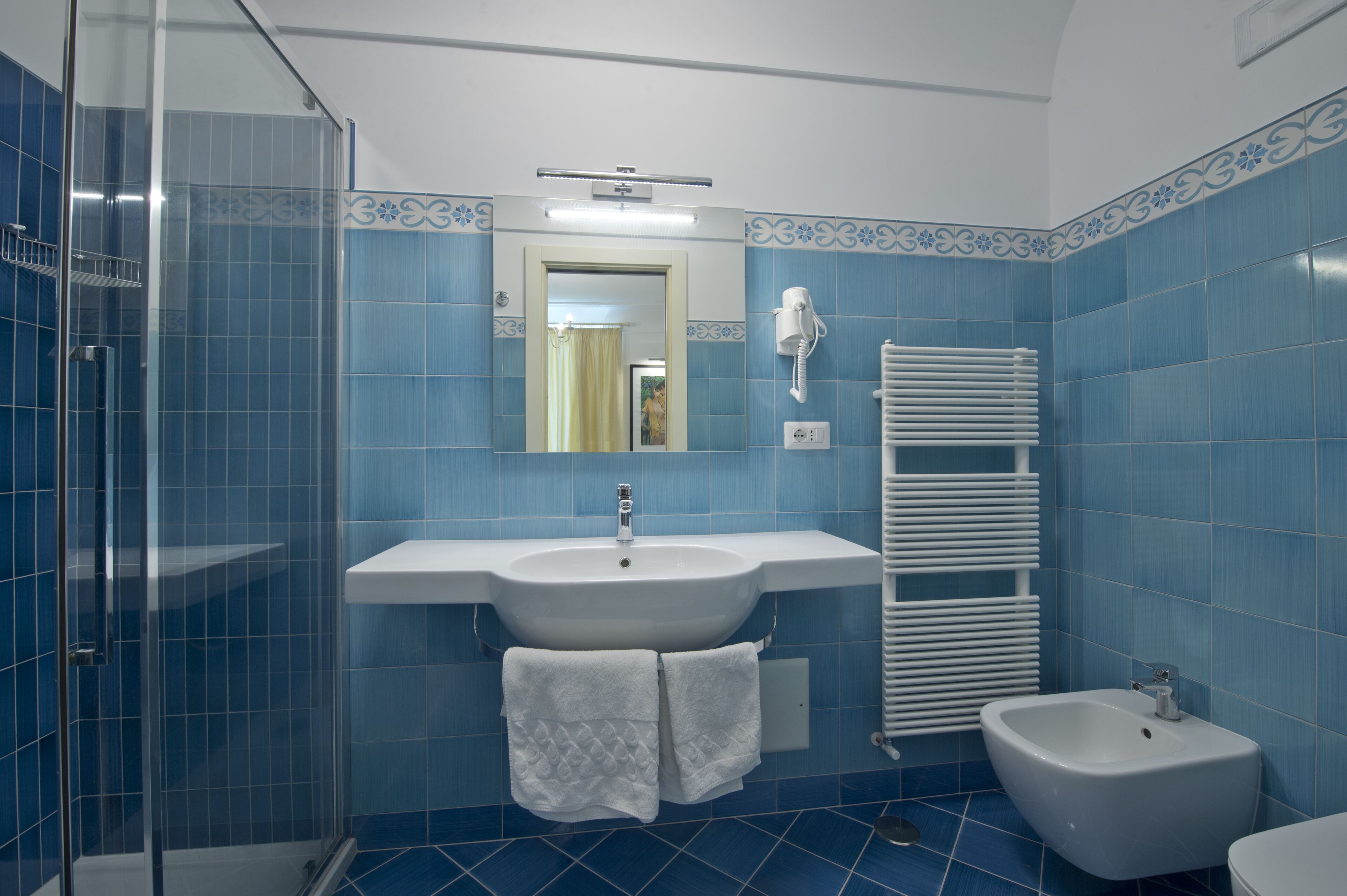 A bathroom with blue tiles, a white sink, a mirror, a towel rack, a shower, and a bidet.
