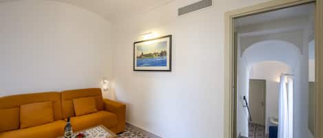 A living room with a sofa, a coffee table with a bottle and fruit, patterned floor tiles, a framed picture on the wall, and a door leading to another room.